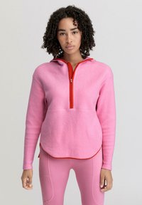 Pullover in pile rosa con colletto zip rosso e cappuccio. Presenta maniche lunghe e orlo inferiore arrotondato. Tessuto testurizzato.