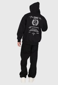 Osoba w czarnej bluzie z kapturem, z białym tekstem i logo streetwear na plecach, ubrana w luźne czarne spodnie i białe sneakersy, na szarym tle.