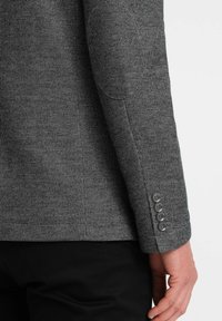 Manga de blazer texturizado gris con parche en el codo, que cuenta con cinco botones oscuros en el puño y un corte entallado. Pantalones negros visibles por debajo.