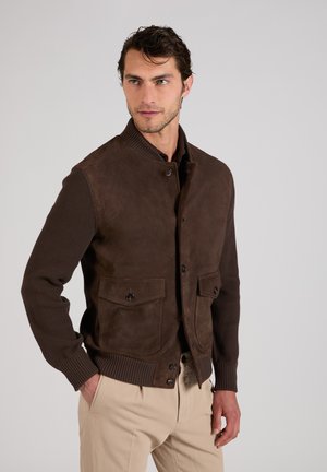Hombre con chaqueta marrón oscuro con mangas de punto y pantalones beige, de pie con la mano izquierda en el bolsillo, mirando ligeramente hacia la derecha.