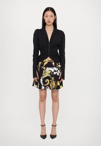 Svart tettsittende blazer med rynket design, med glidelås, kombinert med et mønstret skjørt som har gule og hvite svirvler på svart bakgrunn.