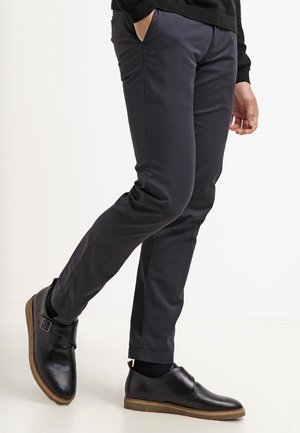 Person iført mørkeblå slim fit bukser og sorte læderbæltesko, stående med det ene ben let løftet.