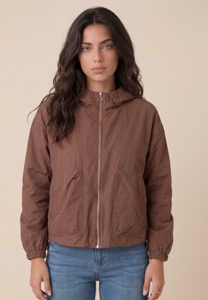 Frau mit langen dunklen Haaren, die eine braune Jacke mit Reißverschluss und elastischen Bündchen sowie blaue Jeans vor einem schlichten beigen Hintergrund trägt.