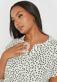 Camicetta bianca con pois neri, maniche corte e scollatura a chiave. Tessuto strutturato con una vestibilità rilassata e dettagli sottili.