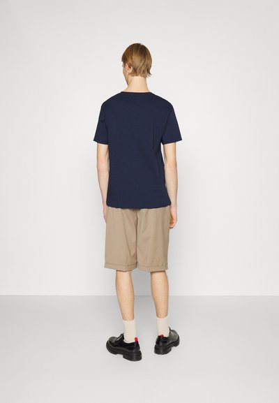 Tricou din bumbac în nuanță navy, cu mâneci scurte, asortat cu șorturi bej. Modelul poartă pantofi negri și șosete crem, văzut din spate.