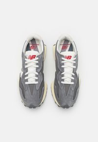 Pár šedých a bílých tenisek New Balance s červeným logem, bílými tkaničkami a béžovými podrážkami, z pohledu shora na bílém pozadí.