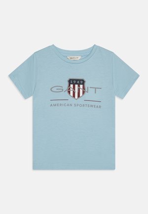 Šviesiai mėlynas medvilninis marškinėliai su trumpomis rankovėmis, su tamsiai mėlynu ir raudonu skydu logotipu ir "GANT AMERICAN SPORTSWEAR" užrašu centre.