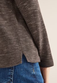 Maglione marrone lavorato a maglia con una finitura testurizzata, caratterizzato da una fessura laterale all'orlo. Abbinato a jeans blu.