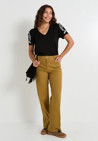 Camiseta negra de cuello en V con bordado floral blanco en las mangas, combinada con pantalones mostaza de cintura alta y zapatos de estampado de leopardo marrón.