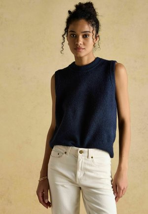 Marineblauer ärmelloser Strickpullover mit geripptem Ausschnitt, kombiniert mit weißen, hoch taillierten Jeans mit goldenen Knopfakzenten.