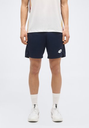 Mann trägt marineblaue Sportshorts, weiße Socken, weiße Sportschuhe und ein weißes Hemd mit Seitenstreifen, steht vor einem einfarbigen Hintergrund.