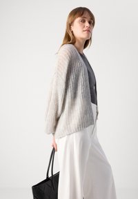 Lichtgrijze, opengehaakte cardigan met lange, brede mouwen, gedragen over een donkere top, in combinatie met witte broeken. Inclusief een zwarte schoudertas.