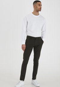 Dunkelgrüne Slim-Fit-Hosen mit flachem vorderen Teil, Gürtelschlaufen und einem einzigen Knopfverschluss, kombiniert mit einem schlichten weißen Langarmshirt und weißen Sneakers.