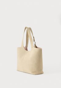 Sac fourre-tout en daim beige avec doubles bretelles, design à coutures courbes et doublure intérieure rose pâle, posé sur un fond blanc.