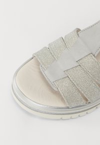 Grå sandaler med brede, krysslagte stropper av glitrende og glatt materiale, med en skinnende sølvsåle og en myk beige foteng.