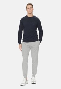 Felpa blu navy con logo "GUESS" in rilievo, abbinata a pantaloni jogger grigio chiaro. Il modello indossa sneakers bianche. Stile casual semplice e sportivo.