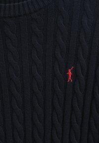 Pull en maille tissée bleu marine avec un motif épais et texturé. Présente une petite figure rouge brodée sur la poitrine, col ras du cou.