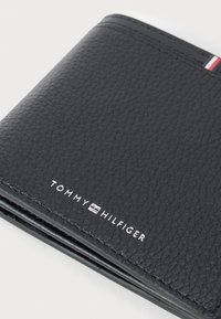 Carteira de couro preto com superfície texturizada, apresentando um logótipo branco e um pequeno detalhe em tecido nas cores vermelho, branco e azul na borda.