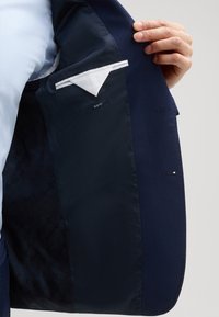 Wnętrze granatowego żakietu z satynową podszewką, metką slim fit, białą kieszonką na chusteczkę oraz teksturowanymi ciemnymi wzorami materiału.