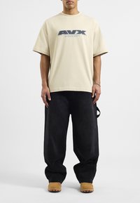 Beżowa koszulka z krótkim rękawem z graficznym logo "AVIX" i napisem "AVIREX DEPT.", zestawiona z luźnymi czarnymi dżinsami i jasnobrązowymi butami wsuwanymi.