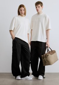 Μπεζ υπερμεγέθη μπλουζάκια με μικρό λογότυπο, μαύροι παντελόνια cargo και λευκά αθλητικά παπούτσια. Κρατιέται μια καφέ τσάντα τύπου duffel με καφέ λεπτομέρειες.