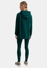 Donna con capelli biondi raccolti in una coda di cavallo, che indossa una felpa verde scuro e leggings abbinati, rivolta di spalle su uno sfondo bianco.