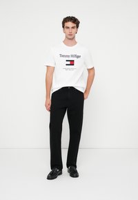 Hvid bomulds T-shirt med "Tommy Hilfiger" logo, parret med sorte bukser. Bukserne har lige ben og klassiske lommer, skoene er sorte læder.