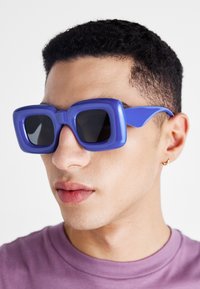 Ochelari de soare mov supradimensionați cu rame p�ătrate, lentile întunecate și un material de plastic neted, purtați de o persoană cu o expresie neutră.