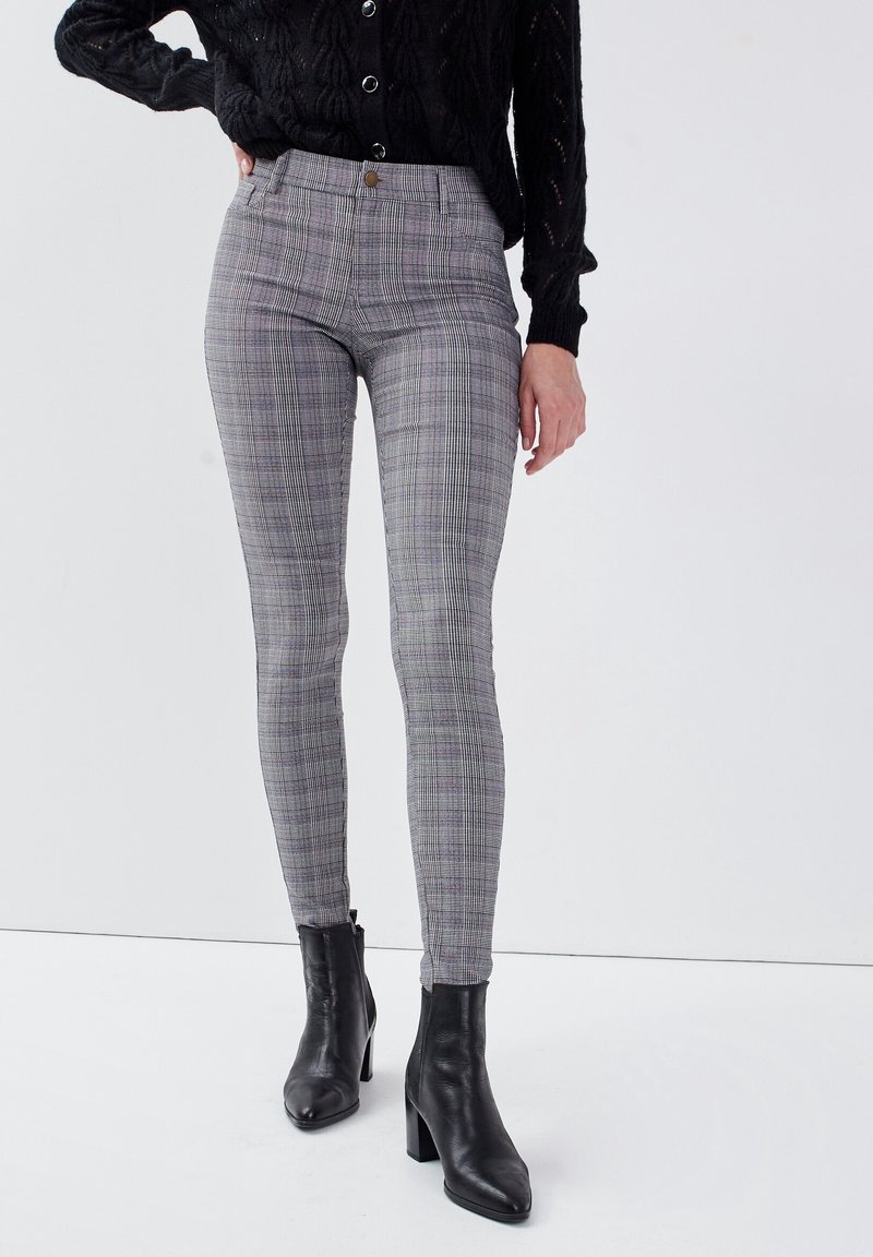 Femme portant un pantalon ajusté à carreaux gris, des bottines noires à talons et un haut noir texturé à manches longues, debout devant un fond blanc.