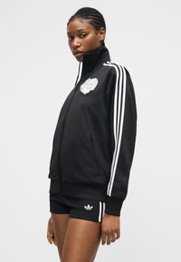 Frau trägt schwarze Adidas-Trainingsjacke und passende Shorts mit weißen Streifen, steht vor einem schlichten hellen Hintergrund.