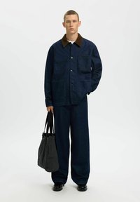 Jeune homme debout face à l'avant, portant une veste en denim bleu foncé et un pantalon large assorti, tenant un grand sac fourre-tout noir dans une main.