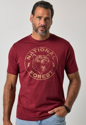 Uomo con barba che indossa una maglietta rossa con la grafica di un orso ruggente e le parole "National Forest,", in piedi con una mano in tasca.
