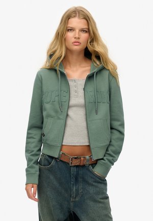 Jonge vrouw met lang blond haar, dragend een groene zip-up hoodie, een grijs cropped henley topje, losse spijkerbroek en een bruine gespen riem met studs.