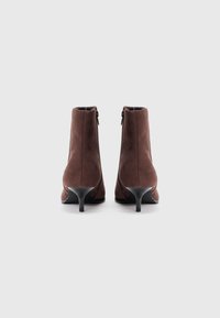 Botas de tornozelo em camurça castanha com um salto fino preto, fechamento com zíper na parte de trás e uma textura suave. Projetadas com uma silhueta ajustada.