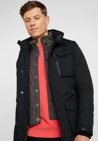 Black outdoor jacket with a detachable quilted liner, featuring multiple pockets, snap closures, and a high collar. Underneath is a red sweatshirt.