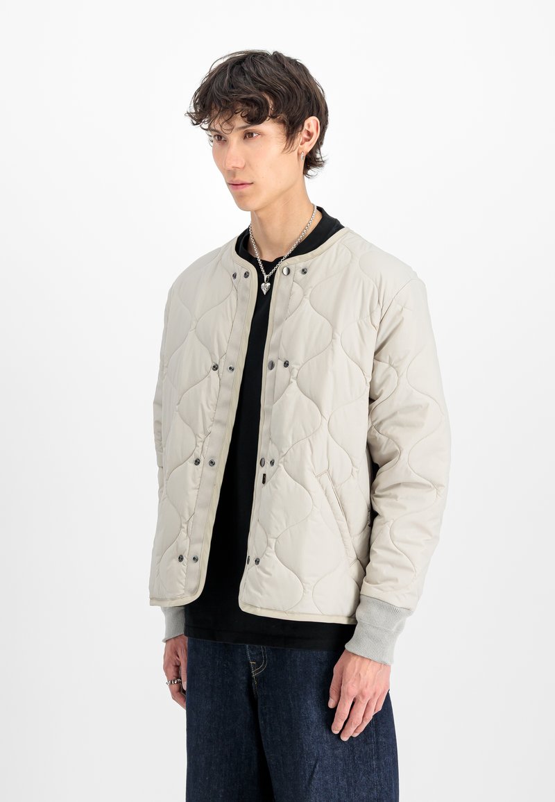 Jauns vīrietis valkā bēšā krāsā šūto jaku pār melnu krekli, tumši zilu džinsu, sudraba kaklarotu ar sirsniņas piekariņu un vairākiem gredzeniem.