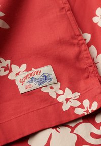 Tela de algodón roja con flores blancas de hibisco y un etiquetado cosido que dice "SUPERDRY BEACH CASUALS" en rojo y azul.
