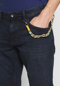 Kleurrijke gevlochten polsband met oranje, blauwe en zwarte patronen, bevestigd aan donker denim jeans, vastgezet met een zilveren clip.
