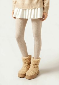 Strikkede beige leggings med et kabelmønster, parret med tanfarvede, fluffy ankelstøvler og en hvid plisseret nederdel. Blød tekstur gennem hele outfits.