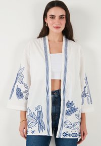 Valge kimono stiilis jakk, millel on sinised tikitud lehe- ja papagoi mustrid, mustriline äär ja vabalt langevad varrukad.