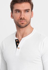 Mann mit kurzen braunen Haaren und Bart, der ein weißes Langarm-Henley-Shirt mit drei beigen Knöpfen und einer schwarzen Knopfleiste darunter trägt.