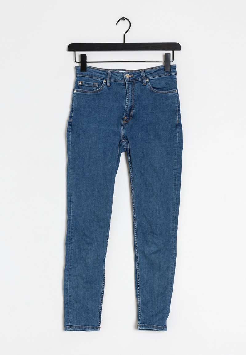 Jean denim bleu slim-fit suspendu sur un cintre noir contre un fond blanc, avec les poches avant et la fermeture à boutons visibles.