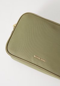 Olīvu zaļas krāsas auduma maisiņš ar teksturētu virsmu, ar zeltītu "Michael Kors" logo un gludām, noapaļotām malām.