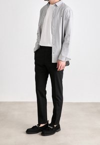 Uomo che indossa una camicia grigia chiaro con bottoni sopra una t-shirt bianca, pantaloni neri slim-fit e mocassini neri con dettaglio a catena, in piedi su un pavimento grigio.