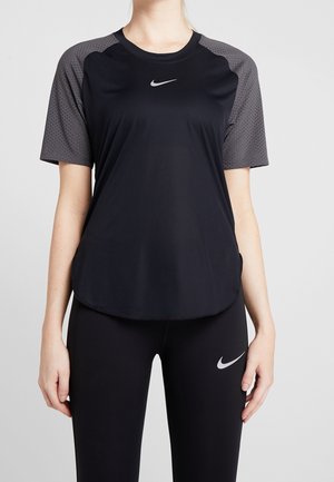 Mujer vestida con camiseta deportiva negra de Nike con mangas grises perforadas y leggings negros con el logo de Nike, de pie frente a un fondo liso.