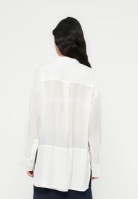 Λευκή μπλούζα με μακριά μανίκια και διαφανή υφή, με πίσω γιακά, κουμπωτά μανίκια και πλαϊνές σχισμές. Λείο ύφασμα μ ε άνετη εφαρμογή.