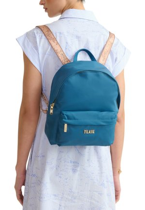 Donna con zaino di nylon blu con cerniere dorate e spallacci beige con motivo a mappa, indossa un vestito azzurro chiaro con stampa a mappa.