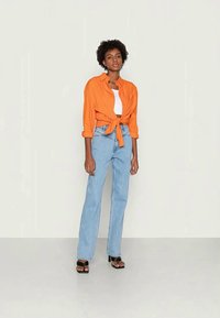 Camisa laranja com laço à frente, top branco, e jeans de cintura alta azul claro. Sandálias de salto pretas. Fundo simples.