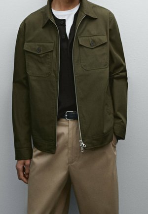 Homme portant une veste zippée vert olive sur une chemise noire boutonnée et un sous-vêtement blanc, associé à un pantalon beige et une ceinture noire.