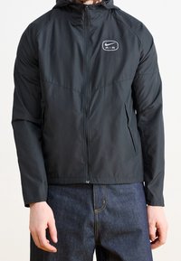 Černá lehká bunda se zipem vpředu, kapucí a logem na prsou. Má hladkou texturu, elastické manžety a boční kapsy.